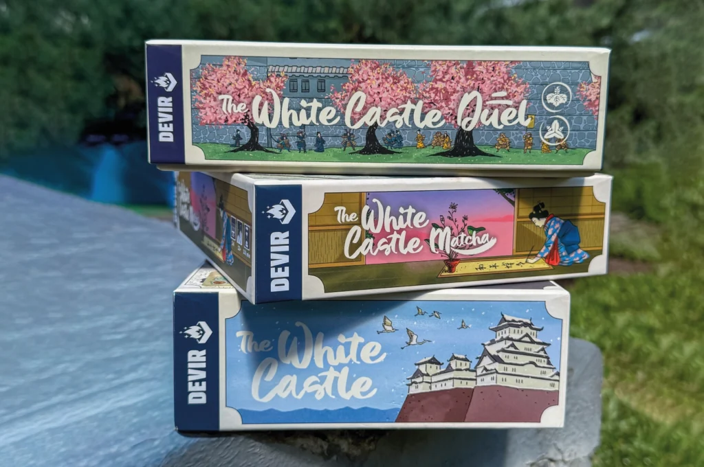 Três caixas da coleção The White Castle da Devir empilhadas ao ar livre, com ilustrações temáticas japonesas.