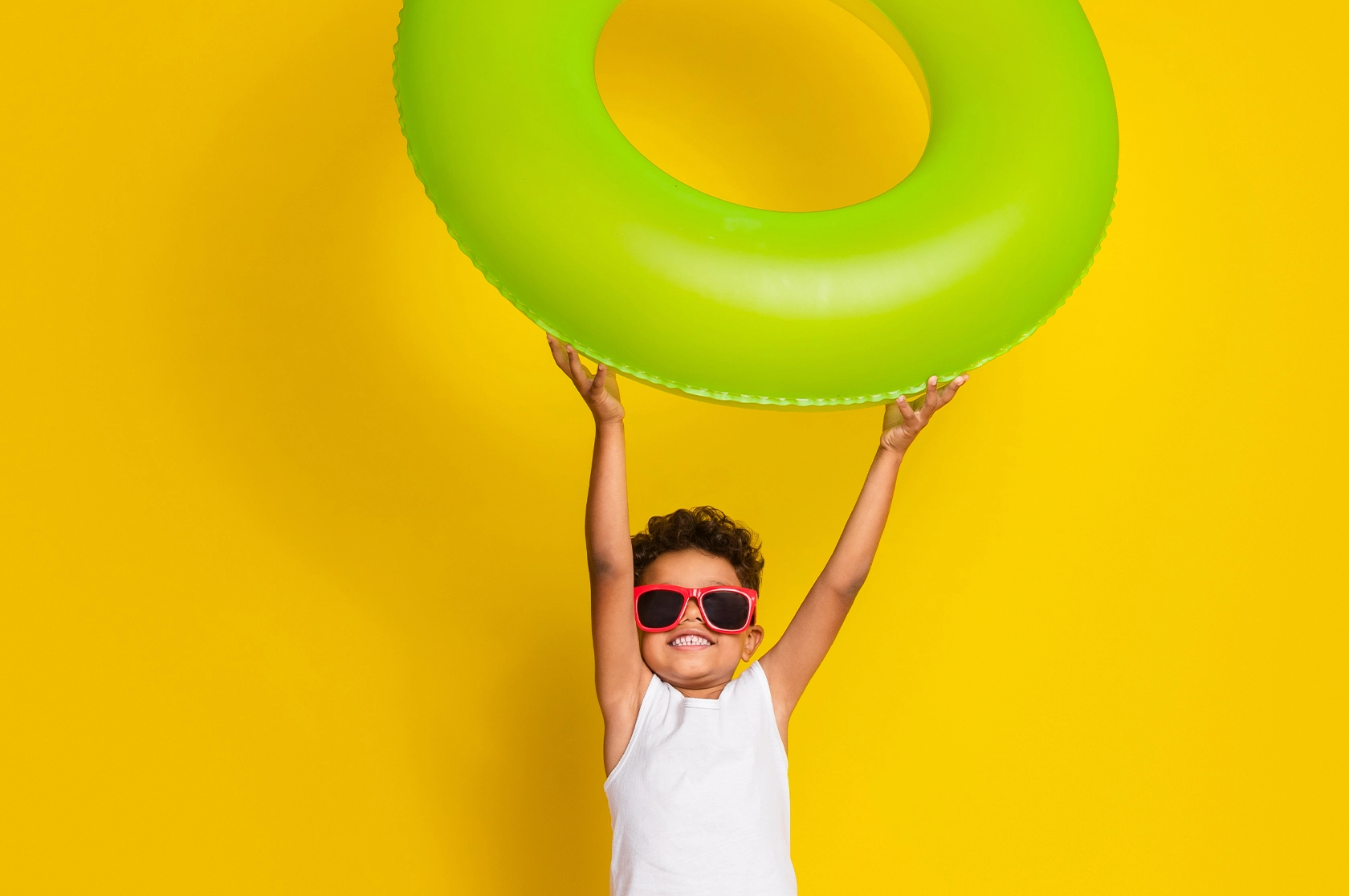 Criança feliz, com óculos de sol vermelhos a segurar numa boia verde, em fundo amarelo