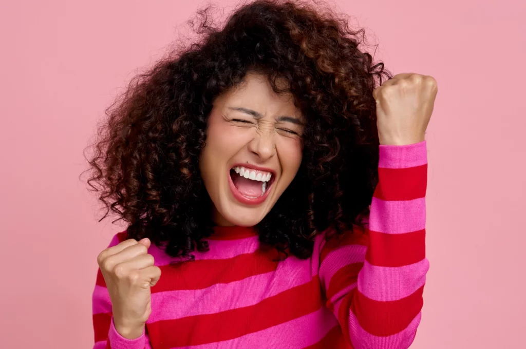 Pessoa em pose de celebração, com os braços levantados e fundo rosa.