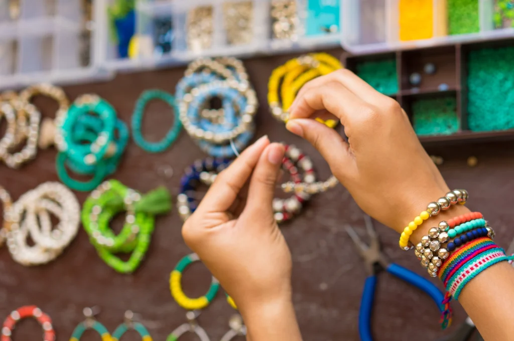Mãos a criar pulseiras artesanais coloridas numa banca de acessórios feitos à mão.